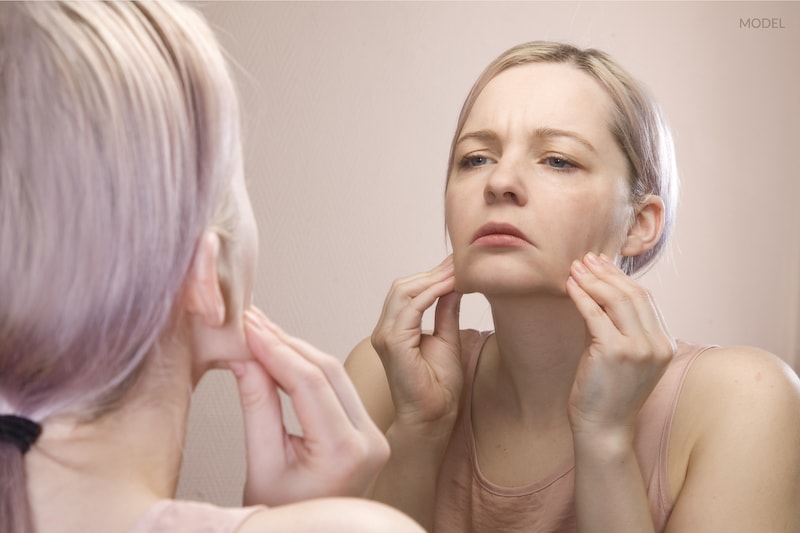 Woman pulling her jowls downward while looking in mirror at her reflection.